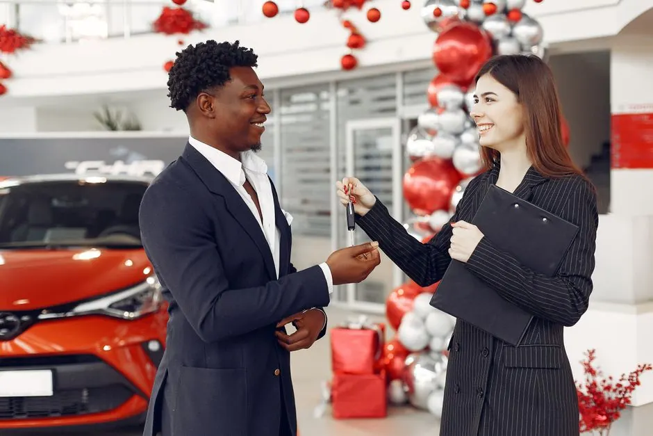 Side view of happy positive female dealer in formal classy wear passing car keys to  excited African American customer in trendy suit in car showroom